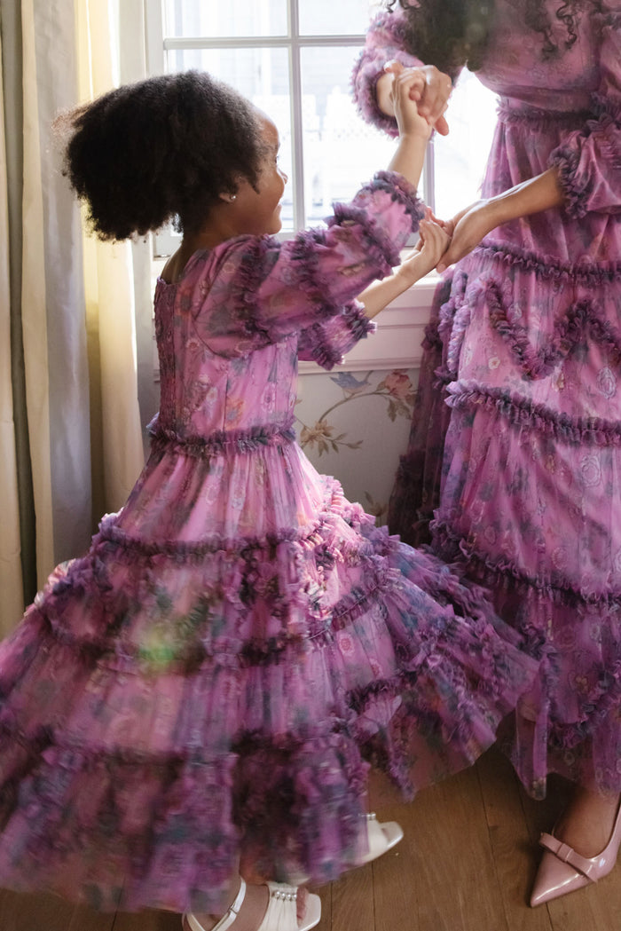 A child and an adult, both in Ivy City Co’s Mini Catherine Dress in Climbing Vine Bloom, hold hands and dance by a sunlit window—their matching floral outfits capturing a joyful moment together.