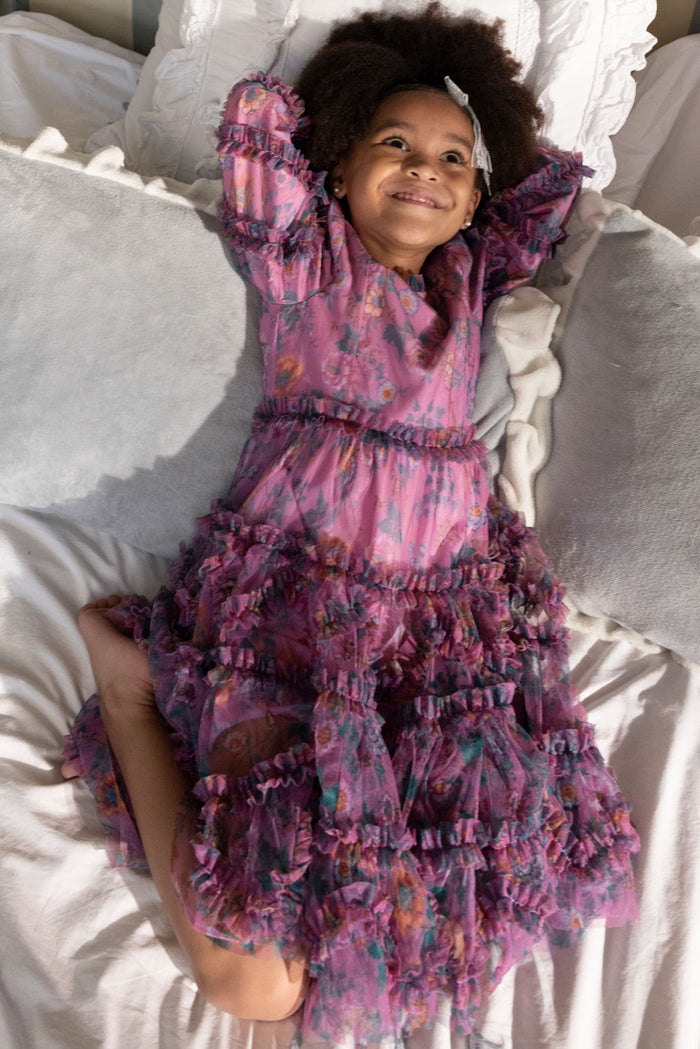 A young child with curly hair, wearing Ivy City Co’s Mini Catherine Dress in Climbing Vine Bloom, lies on a bed with white pillows, smiling and stretching—ideal for capturing matching moments.