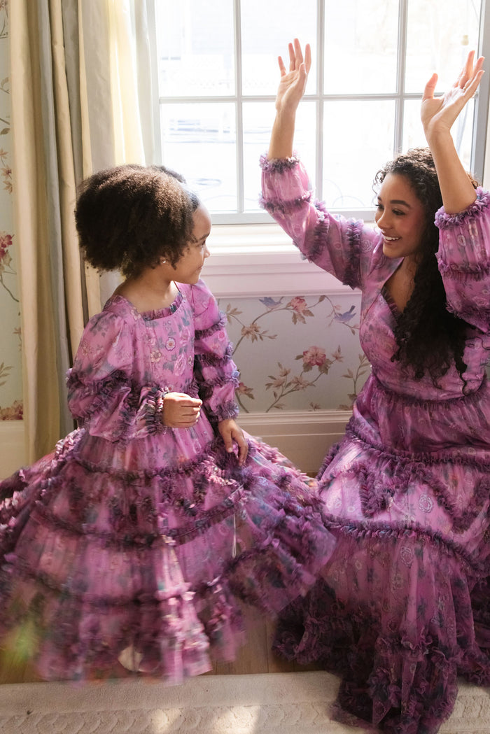 A woman and a young girl joyfully dance indoors by a sunlit window, wearing matching Ivy City Co Mini Catherine Dresses in Climbing Vine Bloom, creating sweet moments against floral wallpaper.