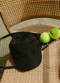 Black Percival nylon cap with PCVL embroidery, resting on a tennis racket and balls on a wicker chair.