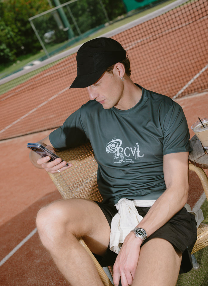 Forest green Percival PCVL printed performance top, worn by a man sitting court-side at a tennis court.