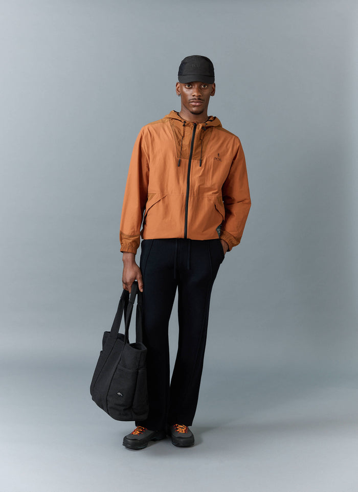 A model wears the Percival PCVL Zip Through Water Resistant Jacket in rust, styled with black trousers, a cap and a tote bag.