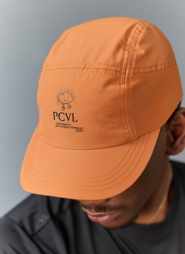 A close-up of a model wearing the rust-coloured PCVL Nylon Cap from Percival, showing the embroidered front logo.
