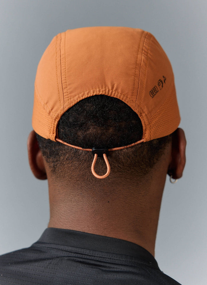 A model wears the Percival PCVL Nylon Cap in rust, viewed from behind to show the adjustable cord and side logo.