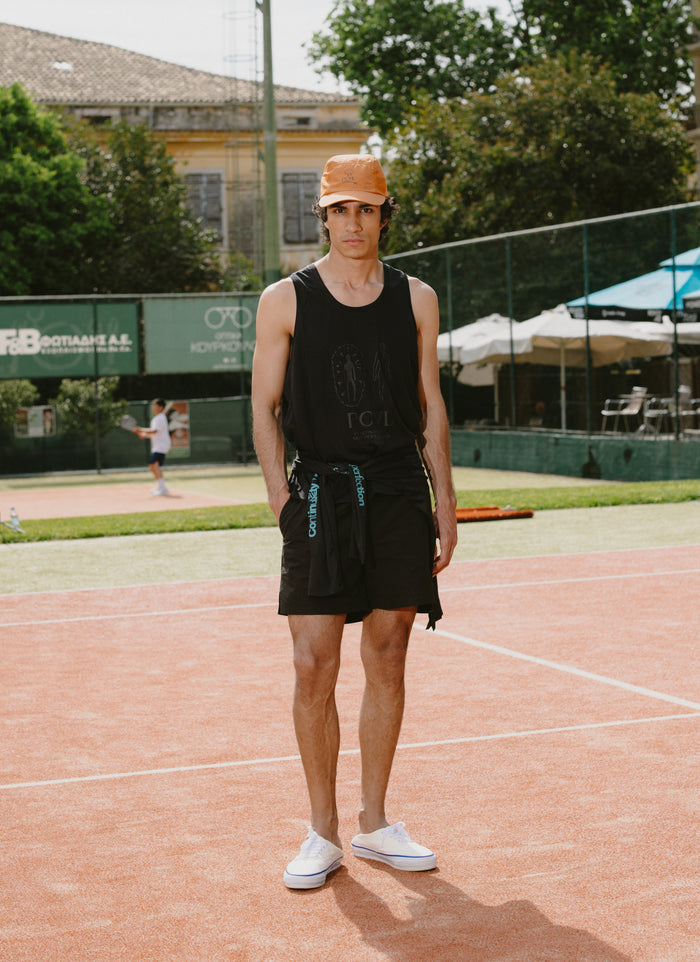 A rust orange Percival nylon cap styled with a black performance vest on a red clay tennis court.