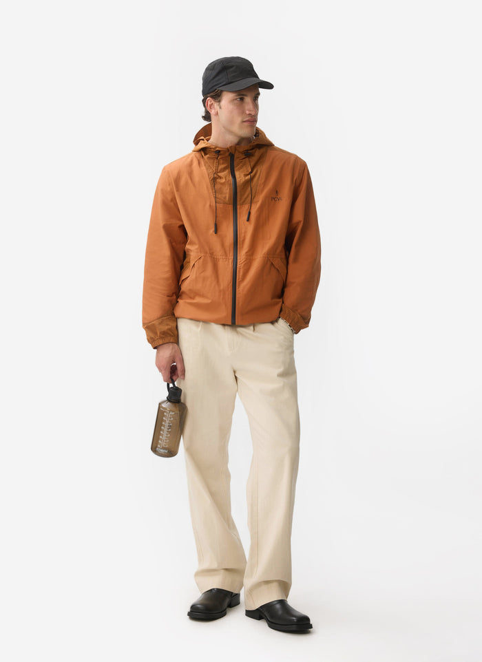 A model in a brown jacket and cream trousers holds the brown PCVL Water Bottle from Percival.