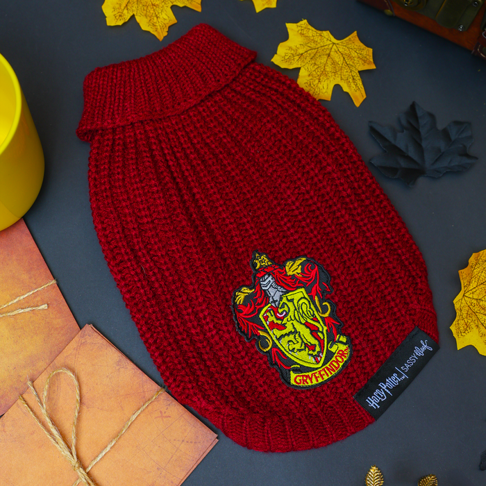 The Sassy Woof Pet Cable Knit Sweater - Harry Potter™ Gryffindor, featuring a vibrant red knit and the iconic Gryffindor crest, is artfully displayed among autumn leaves, a yellow cup, a chest, and wrapped gifts on a dark background—capturing the cozy charm of this magical dog sweater for your furry companion.