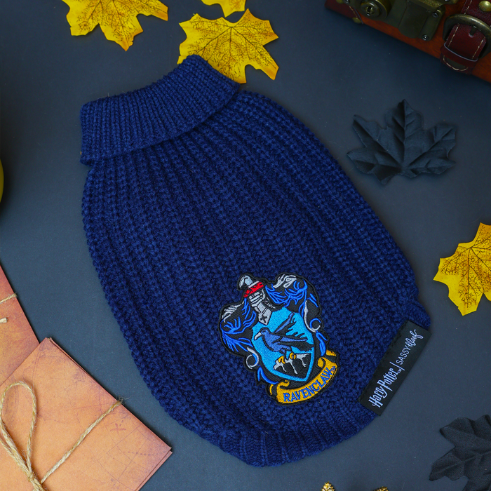 A Sassy Woof Pet Cable Knit Sweater - Harry Potter™ Ravenclaw, featuring a blue cable knit design with the iconic Ravenclaw crest patch, rests on a dark surface surrounded by yellow leaves, a brown book, and a small chest—making it the ideal cozy sweater for your dog during cool autumn days.