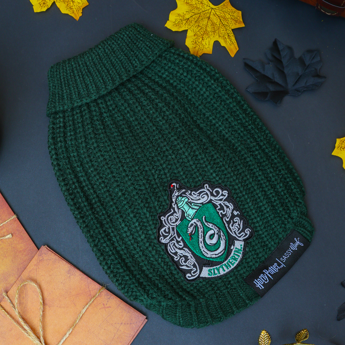 A green Pet Cable Knit Sweater - Harry Potter™ Slytherin by Sassy Woof, featuring a Slytherin crest patch, is displayed on a dark surface with yellow and black paper leaves and brown envelopes—a cozy sweater ideal for any fan of Slytherin dog apparel.