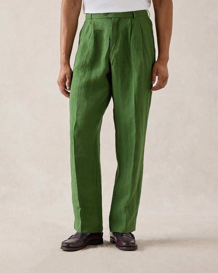 A model wearing Percival forest green pleated linen trousers with belt loops, paired with a white t-shirt and loafers.