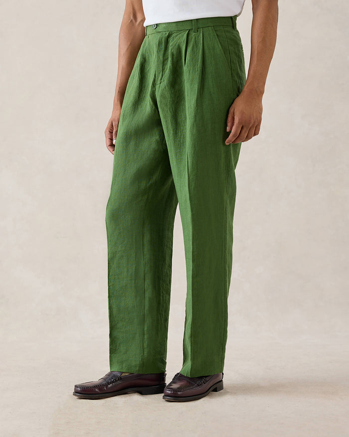 A model wears forest green Percival pleated linen trousers with belt loops and a white t-shirt in a studio shot.