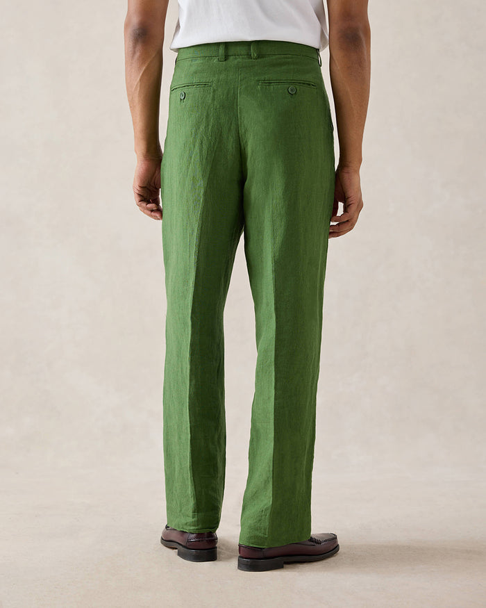 A rear view of a model wearing Percival forest green pleated linen trousers with belt loops and back welt pockets.