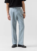 A model wears the Percival Pleated Tailored Trousers in light blue linen with a black top and black loafers.