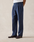 A model wears navy blue Percival tailored linen trousers with belt loops and pleats, styled with a white t-shirt and loafers.