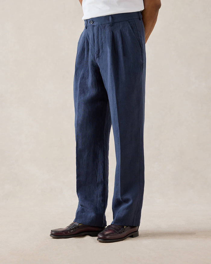 A model wears navy blue Percival tailored linen trousers with belt loops and pleats, styled with a white t-shirt and loafers.