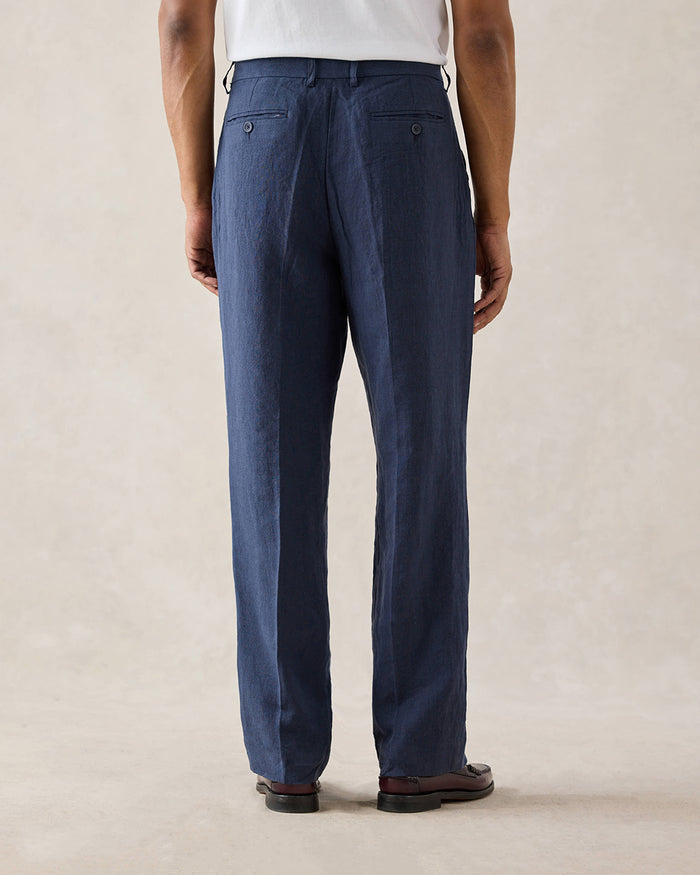 A rear view of a model wearing Percival navy pleated linen trousers with belt loops and back welt pockets.