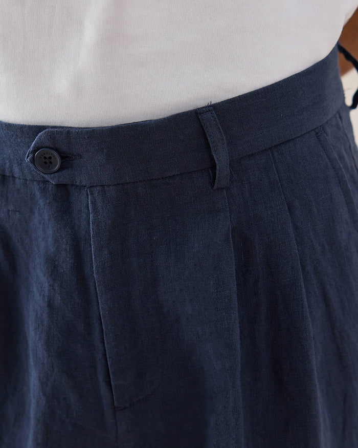 Close-up of the waistband and belt loops on navy Percival pleated linen trousers, worn with a white t-shirt.