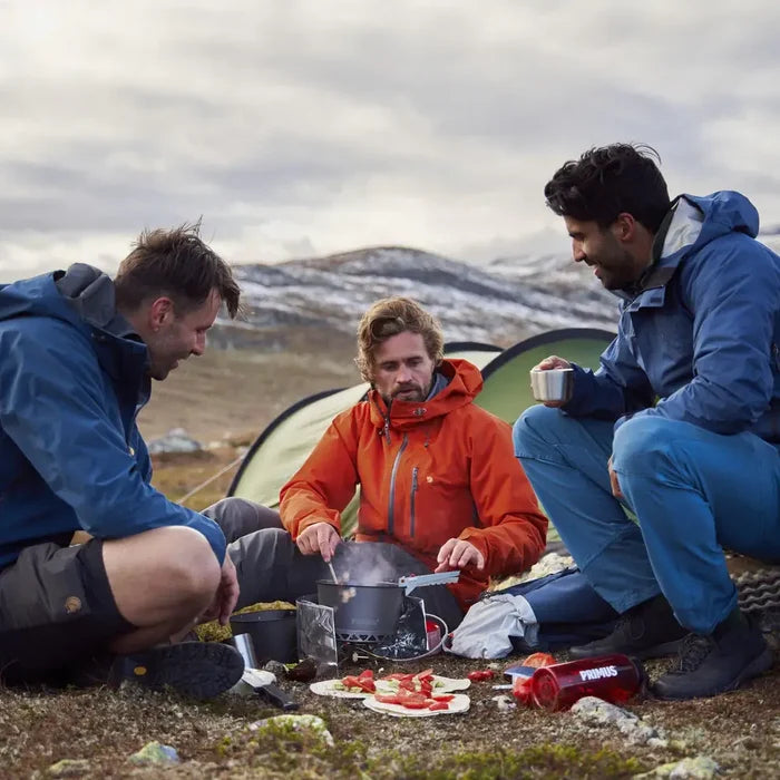 Three people camping in a mountainous area, cooking outdoors using a Primus Gravity Camping Gas Stove.