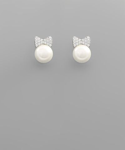 Pearl stud earrings with crystal ribbon bow detail in gold and rhodium finishes displayed on a gray background from The Pink Doxie Boutique.