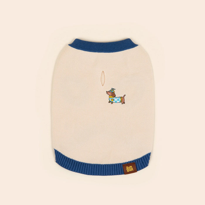 The Sunbean Sassy Woof x Sunbean Dog Tee is a cream-colored pet sweater with blue trim, featuring a small embroidered dog wearing a hat and scarf on the back, and a brown patch label sewn near the bottom edge.
