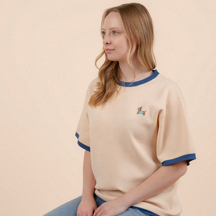 A woman with long, light brown hair sits sideways against a plain light background, wearing the Sunbean Sassy Woof x Sunbean Human Tee in beige with blue trim and a small embroidered dachshund on the chest.