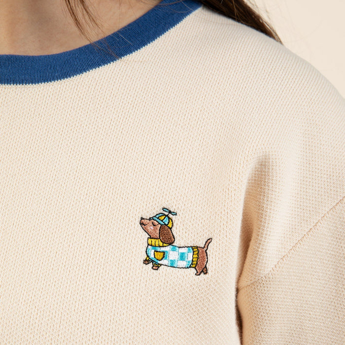 A close-up of the cream-colored Sassy Woof x Sunbean Human Tee by Sunbean, accented with blue trim and featuring an embroidered dachshund sporting a blue hat and a blue-and-yellow checkered sweater—perfect for those who love the matching dog and owner tee trend.