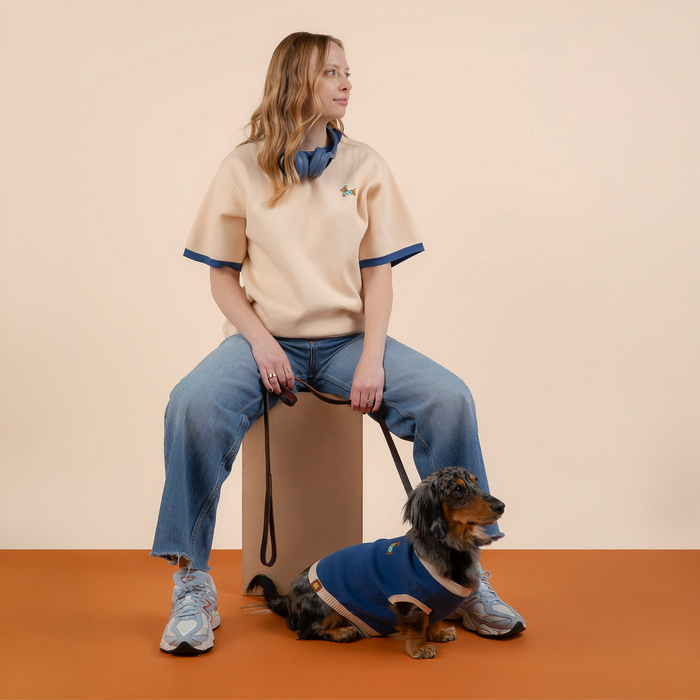 Dressed in a Sunbean Sassy Woof x Sunbean Human Tee, a person in casual attire sits on a block, holding a dog leash. In front of them, a small dog sporting a blue vest sits on the orange floor against a beige background, perfectly matching their owner's tee.
