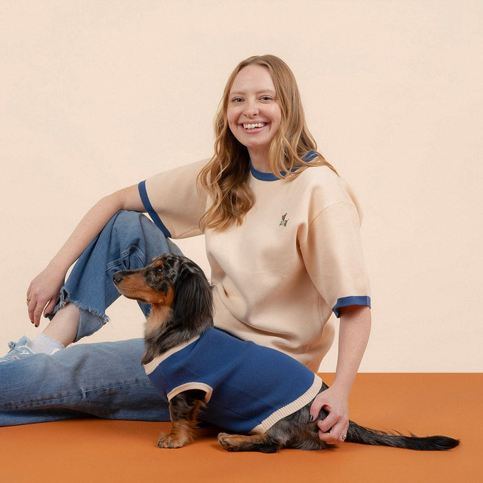 A smiling person wearing the Sunbean Sassy Woof x Sunbean Human Tee in beige and jeans sits on the orange floor beside a small dog in a matching blue sweater, perfectly capturing the adorable Sunbean style. The background is plain beige.