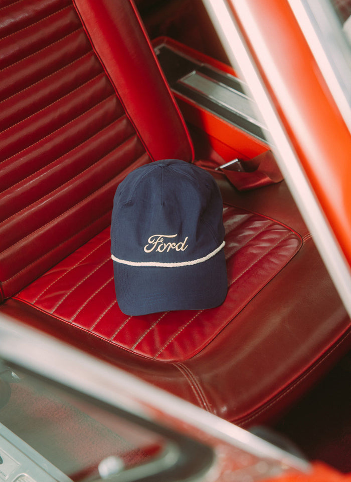 A navy Percival x Ford trucker cap with ecru embroidery sitting on the red leather seat of a classic Ford Mustang.