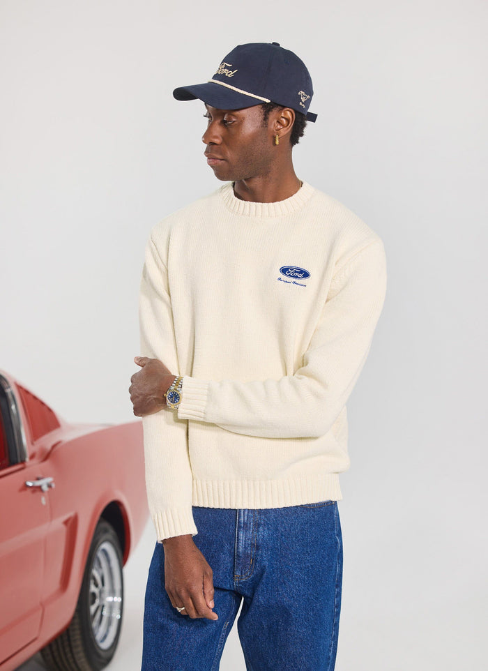 A man models the Percival x Ford ecru jumper and navy cap, standing next to a red car.