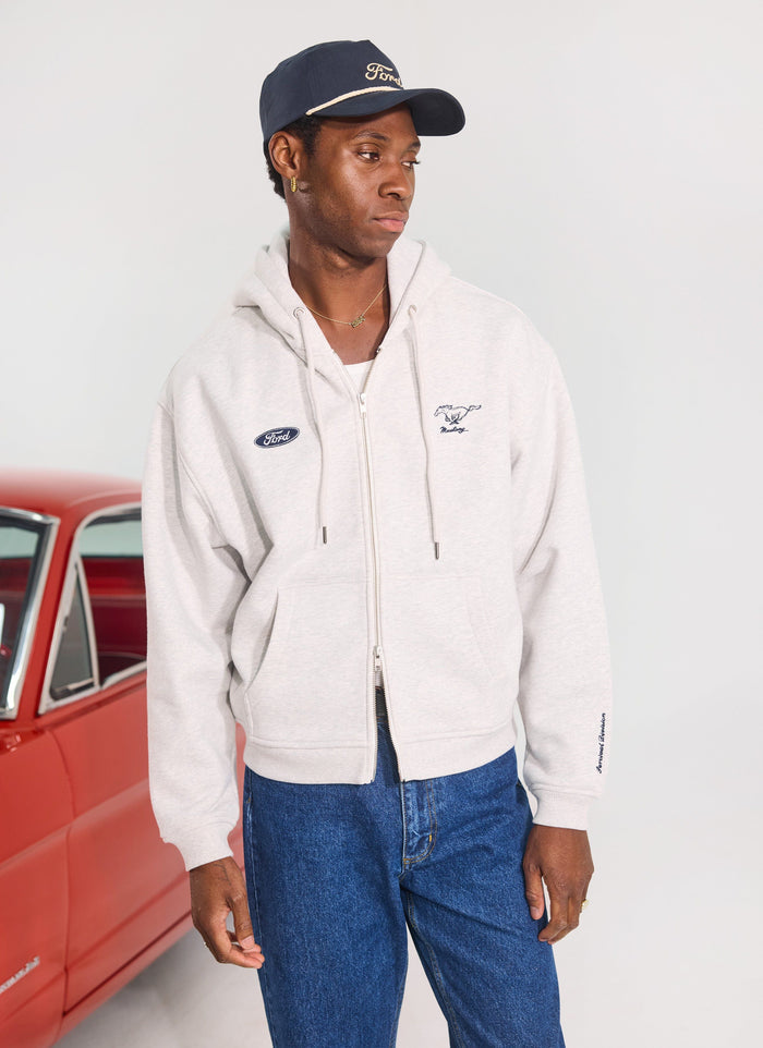 Model wears the Percival x Ford grey zip-through hoodie and navy trucker cap, standing by a vintage red car.