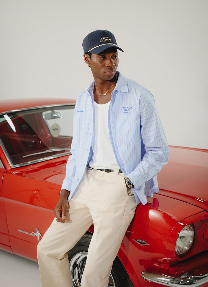 A model wears the blue pinstripe Percival x Ford Pit Crew shirt and a Ford cap while leaning against a classic red Mustang.