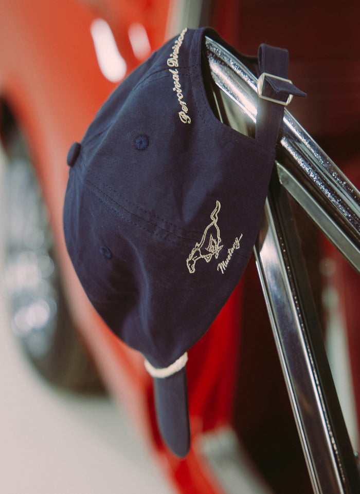 A close-up of the navy Percival x Ford trucker cap showing the 'Mustang' and 'Percival Division' embroidery detail.