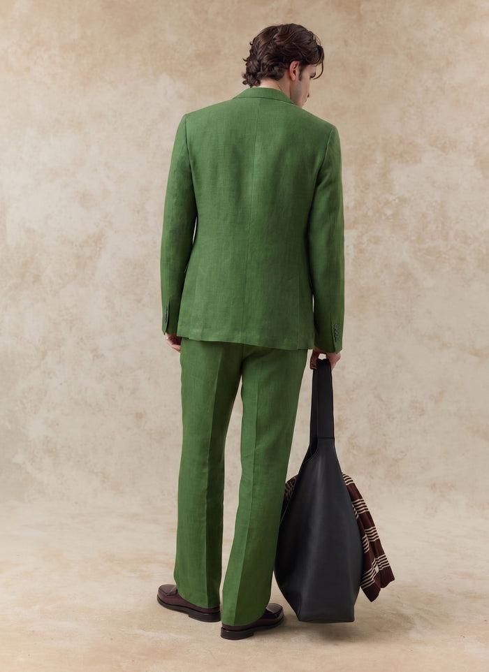 Rear view of a model in the Percival tailored linen trousers and matching blazer in forest green, holding a black bag.