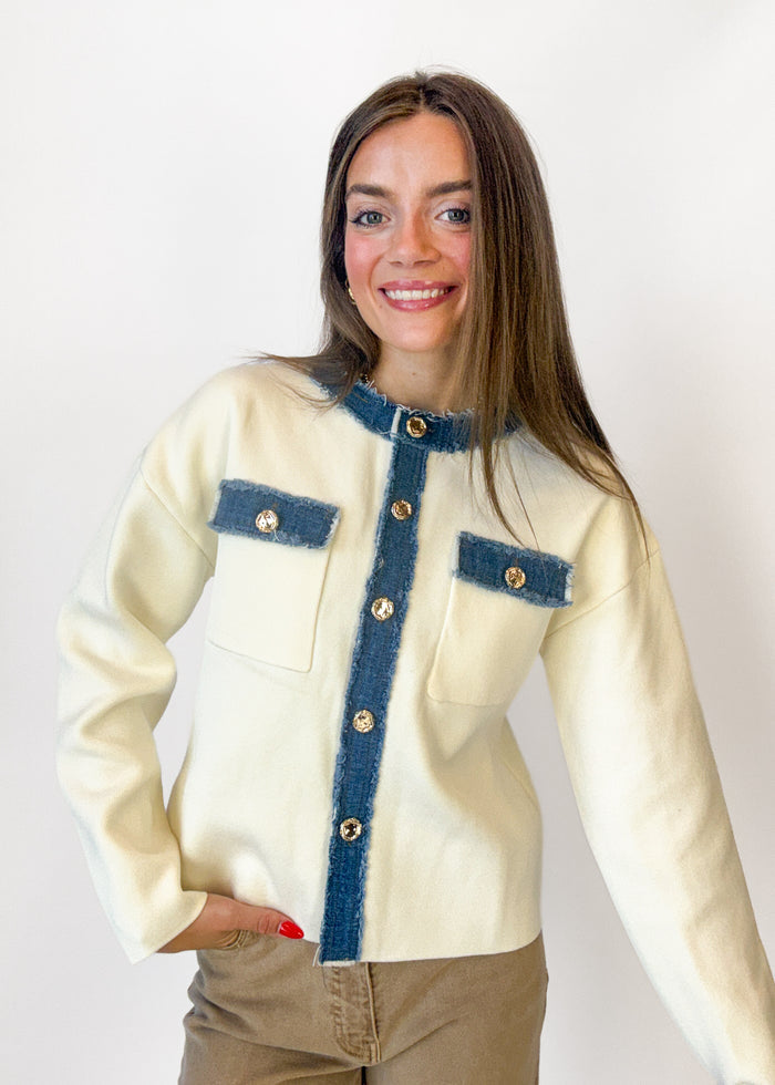 A woman wearing a cream-colored jacket with dark blue denim-style trim and gold-tone buttons, paired with brown pants, photographed against a plain white background.