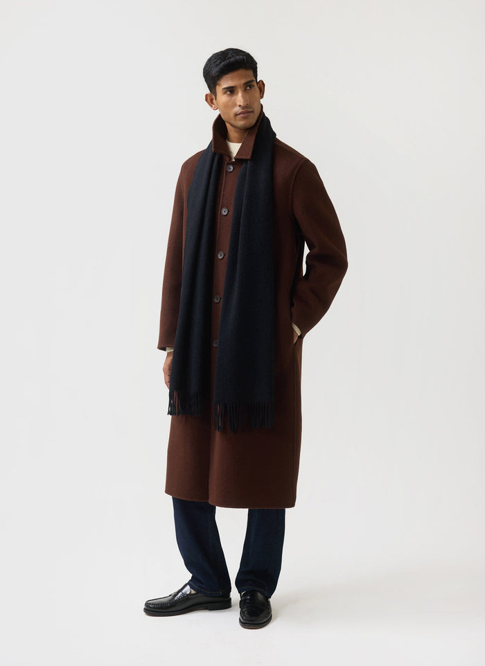 A model wears the black wool Tolkein Scarf from Percival, styled with a long brown coat against a white studio background.